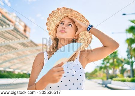 Young beautiful caucasian woman with blond hair smiling happy outdoors using handfan on a hot summer day