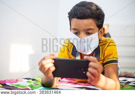 Young Boy Wearing Mask Lying Down On A Bed In A Relaxed Pose, Playing At His Smart Phone. Kid Stayin