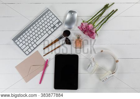 Top view of a tablet computer, a keyboard, a computer mouse, headphones, an envelope, brushes, a perfume, with pink roses arranged on a plain white background