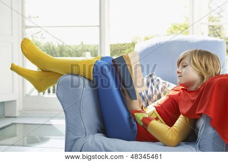 Vista laterale di un giovane ragazzo rilassato in costume da super eroe leggendo il libro sulla poltrona