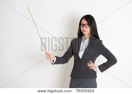 Asian female teacher angry and holding a stick, standing on plain background.