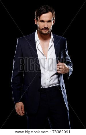 Handsome suave young man in a stylish tailored suit posing with his jacket unbuttoned giving the camera a seductive look on a dark background