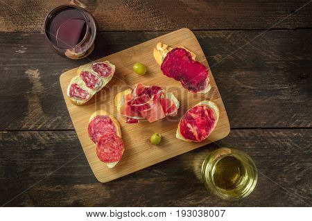 An overhead photo of embutidos tapas, sandwiches with jamon, salchichon, lomo, and other Spanish sausages, with glasses of wine and green olives, on a dark background with a place for text
