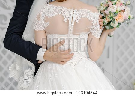 Groom in stylish suit gently hugging beautiful bride with white veil by the waist at the wedding ceremony the back view