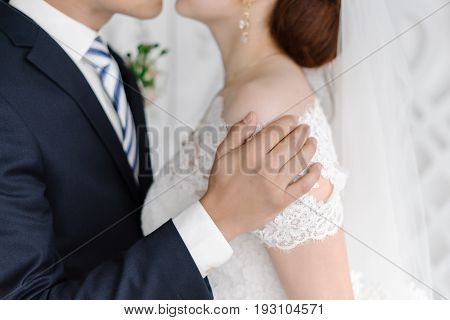 Handsome groom in dark grey suit gently holding beautiful bride with white veil by the shoulders and kissing her at the wedding ceremony