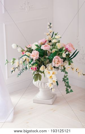 A bouquet of peonies and roses in a clay white jug on the floor at the threshold of the French window. Bright interior.