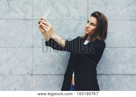 Portrait young businesswomen in black suit using smartphone isolated on background concrete gray wall mockup pretty hipster manager holding mobile gadget girl making selfie photo connect
