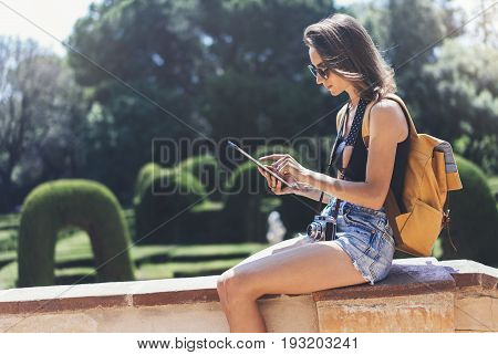 Hipster young girl with bright backpack and vintage camera using tablet or holding gadget planning travel plan. View tourist traveler with sunglasses on background Parc Laberint Barcelona. Mockup for text message