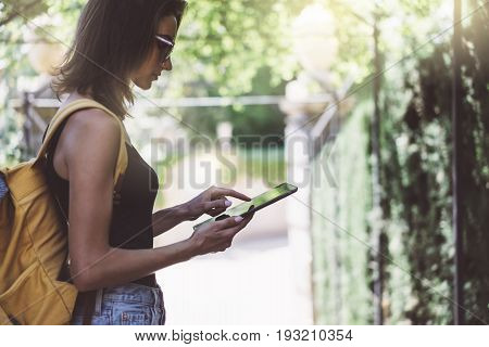 Hipster young girl with bright backpack and vintage camera using tablet or holding gadget planning travel plan. View tourist traveler with sunglasses on background Parc Laberint Barcelona. Mock up for text message