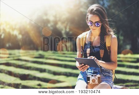 Hipster young girl with bright backpack and vintage camera using tablet or holding gadget planning travel plan. View tourist traveler with sunglasses on background Parc Laberint Barcelona. Mock up for text message