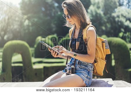 Hipster young girl with bright backpack and vintage camera using tablet or holding gadget planning travel plan. View tourist traveler on background Parc Laberint Barcelona. Mock up for text message