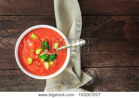 A bowl of gazpacho soup with slices of tomato, cucumber, and onion, with parsley leaves, shot from above on a dark rustic texture with a place for text