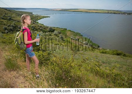 Girl with a backpack standing on the background of the lake and drinking water, sunny day