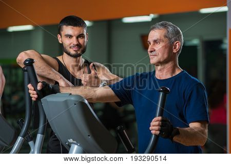 Senior man working with personal trainer in gym. Male adult exercising on elliptical machine with assistance of fitness coach. Healthy lifestyle, fitness and sports concept.