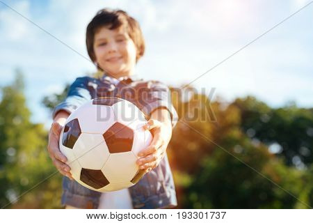 Play with me. Adorable enthusiastic charming boy having an active day while enjoying his hobby and playing football in thee park