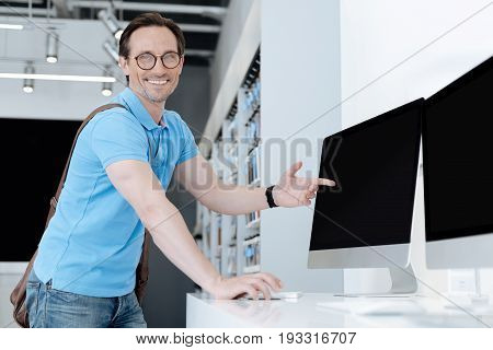 Wrap this one up please. Very excited male customer standing at a display and grinning broadly while shopping for a new computer and pointing his finger at a screen of a mockup gadget.