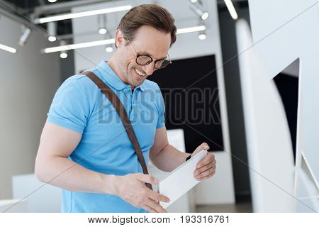 So happy to find it. Side view on a male customer wearing polo holding an information plate and beaming while shopping for a new gadget at electronics shop.