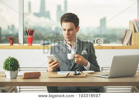 Attractive european businessman using cellphone and drinking coffee at desk with lapop notepad plant and other items. Blurry city background