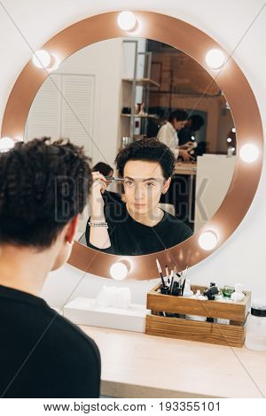 Attractive woman primp at the mirror with lamps in the beauty studio. A happy satisfied customer of make-up and hairstyle service.