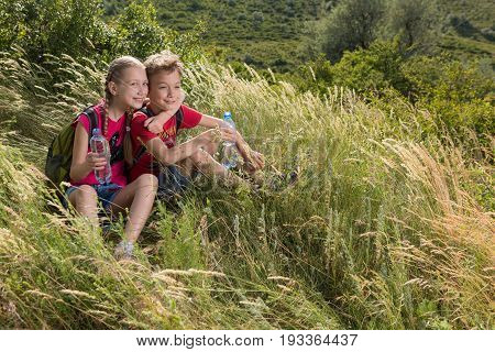Boy and girl are sitting in the tall grass and holding in their hands bottles with water, travel, rest on the glade