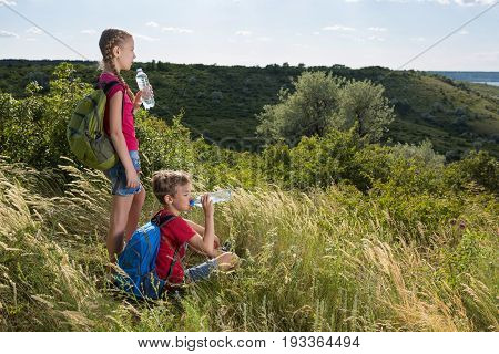 The girl is standing, and the boy is sitting in a clearing with a tall grass and drinking water, traveling, resting on the clearing