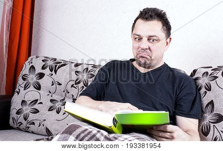 Unshaven Man Sits On A Sofa And Contemptuously Looking At The Book.