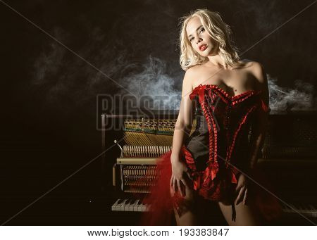 beautiful blonde musician in red corset standing near the piano. Vintage style beautiful woman.. Old fashioned makeup and retro finger wave hairstyle.