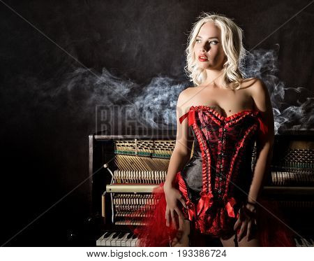 beautiful blonde musician in red corset standing near the piano. Vintage style beautiful woman.. Old fashioned makeup and retro finger wave hairstyle.