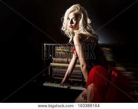 beautiful blonde musician in red corset standing near the piano. Vintage style beautiful woman.. Old fashioned makeup and retro finger wave hairstyle.