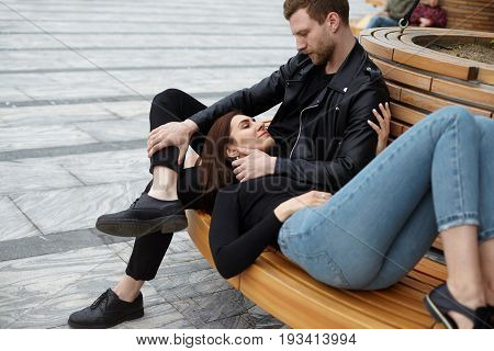 Cute couple cuddling on bench sharing happy moments of love and tenderness. Beautiful girl wearing jeans lying on lap of her attractive bearded boyfriend in leather jacket who is touching her face