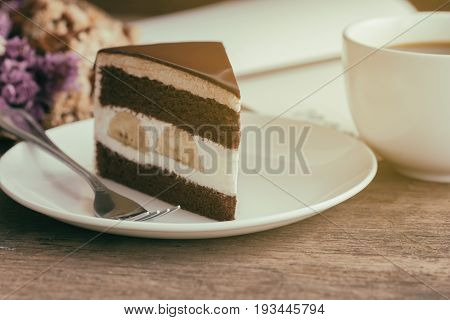 Banana chocolate cake on white plate served with black coffee. Chocolate cake layered with whipped cream and banana look so delicious. Enjoy eating at cafe restaurant with chocolate cake and coffee. Triangle slice piece of chocolate cake.