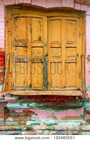 Old sun blind with partially flown paint of yellow color. The window opening is slightly bent. A rusty window sill and a wall of the house put from a brick with partially flown plaster.