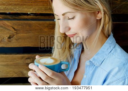 Thoughtful And Pretty Blonde Hair Girl Holding A Cup Of Coffee In Hands. Wood Bacjground In Cafe