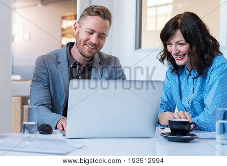 Two smiling work colleagues talking and working together on a laptop while sitting at a table in a meeting room in a modern office