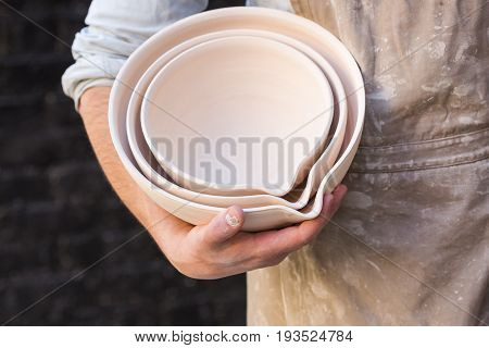 potter, utensils, ceramics art concept - male hands of ceramist holding some beautiful bowls, cute finished clay products and craftsman in the apron on background of dark gray brick wall