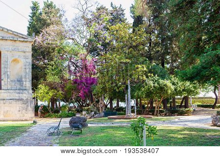 Beautiful green park on Corfu island in Greece. Bench, decorations and a man working on background.