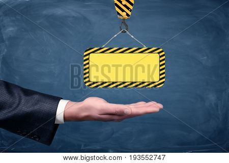 A blank yellow striped construction sign hanging from a crane hook above businessmen's palm on chalkboard background. Development business. Construction works. Building.
