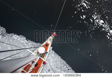 View from the mast on the deck of a ship icebreaker with a team of people. The vessel icebreaker comes out of the water with pieces of ice. A man in red winter clothes is standing on the bow of the ship.