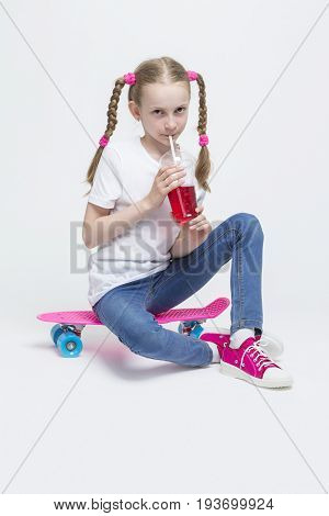 Kids Concepts. Portrait of Little Caucasian Blond Girl with Long Pigtails Posing With Pink Pennyboard and Drinking Red Juice with Straw. Against White. Vertical Image Composition