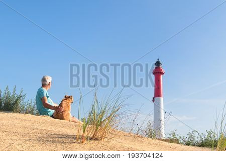Man and his dog sitting on dune and looking to Lighthouse red and white at the French west beach