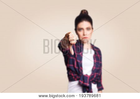 Dislike. Young unhappy upset girl with casual style and bun hair thumbs down her finger, on beige blank wall with copy space looking at camera with toothy smile. focus on finger.