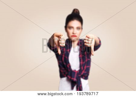 Dislike. Young unhappy upset girl with casual style and bun hair thumbs down her finger, on beige blank wall with copy space looking at camera with toothy smile. focus on finger.