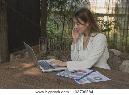 Asian smile beautiful girl sitting outdoor office using smart phone and laptop in nature. Work space with work sheet of business business and lifestyle conceptcopy space