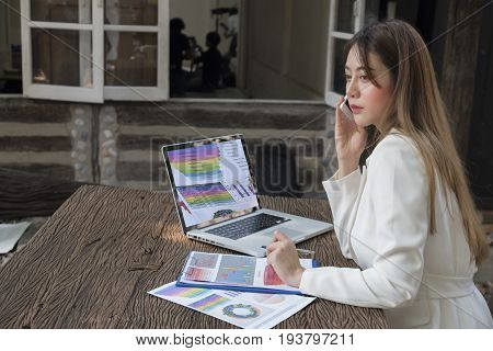 Asian smile beautiful girl sitting outdoor office using smart phone and laptop in nature. Work space with work sheet of business business and lifestyle conceptcopy space