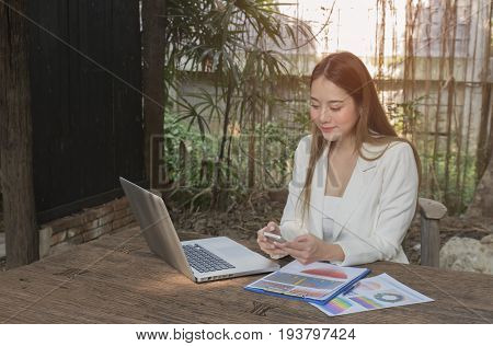 Asian smile beautiful girl sitting outdoor office using smart phone and laptop in nature. Work space with work sheet of business business and lifestyle conceptcopy space