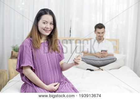 Asian Pregnant Woman Holding Tablet And Husband Behind At Bedroom Together 2 Person Pregnant Woman W