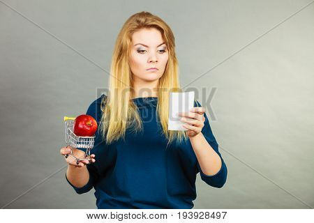 Worried woman holding shopping basket with fruits looking at bill receipt being scared of huge prices