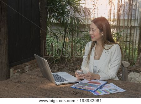 Asian smile beautiful girl sitting outdoor office using smart phone and laptop in nature. Work space with work sheet of business business and lifestyle conceptcopy space
