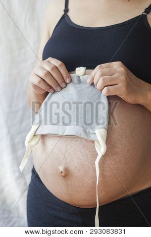 Close Up View Of Nacked Belly Of Pregnant Woman Holding Baby Boy Hat. Pregnant Woman Is Holding A Sm