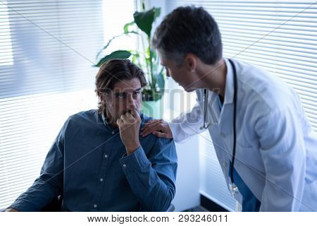 Side view of mature Caucasian female doctor talking with disabled Caucasian patient in wheelchair in clinic at hospital. She tries to make him feel better by putting hand on his shoulder.